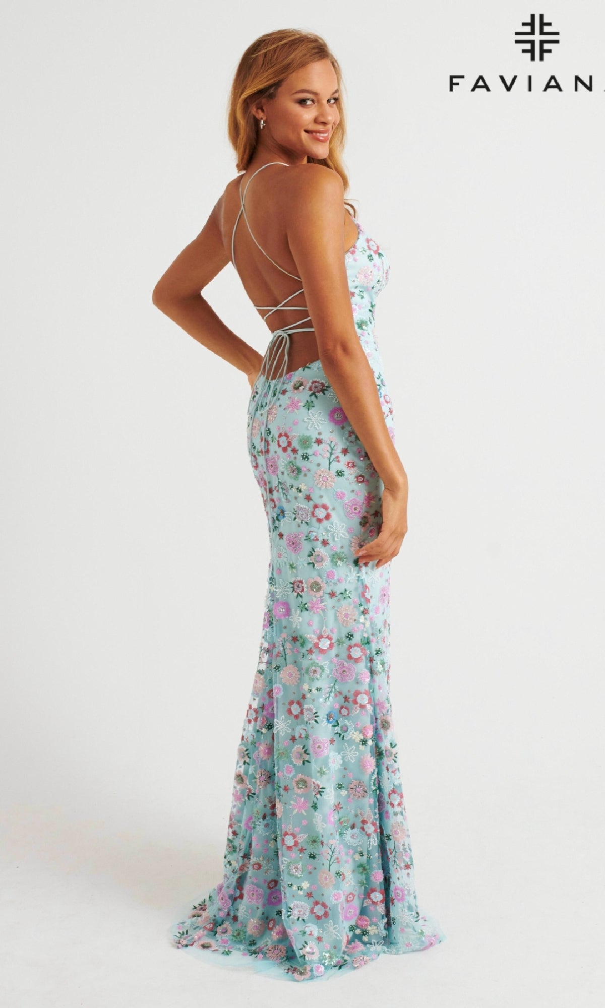 A woman poses sideways in the Faviana 11262 long prom dress—light blue, floral-patterned, floor-length, with an open back and crisscross straps. The brand name “FAVIANA” appears in the top right corner.
